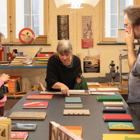 Twee bezoekers krijgen uitleg in het boekbindatelier bij een tafel vol boeken met verschillende soorten boekbanden